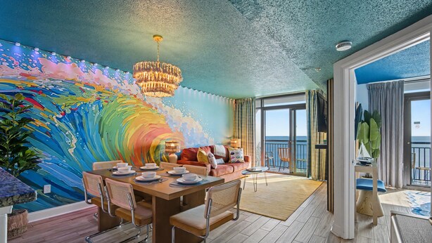 Une pièce comprenant une table à manger dressée pour quatre personnes, un canapé, une grande fenêtre offrant une vue dégagée et une fresque murale aux couleurs vives représentant des vagues.