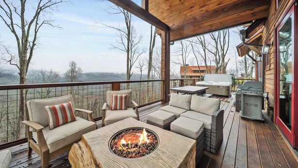 Una terraza de madera con mobiliario exterior, un hogar de fuego y vistas a árboles y edificios.