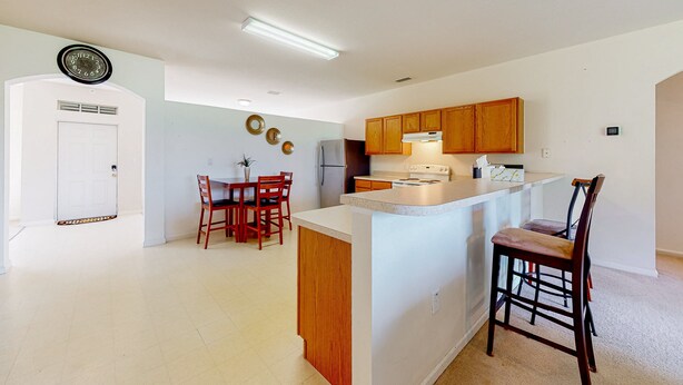 A modern kitchen with wooden cabinets, a white countertop, and a dining area with red chairs. - Palm Bay Cove | House in Palm Bay (Palm Bay)