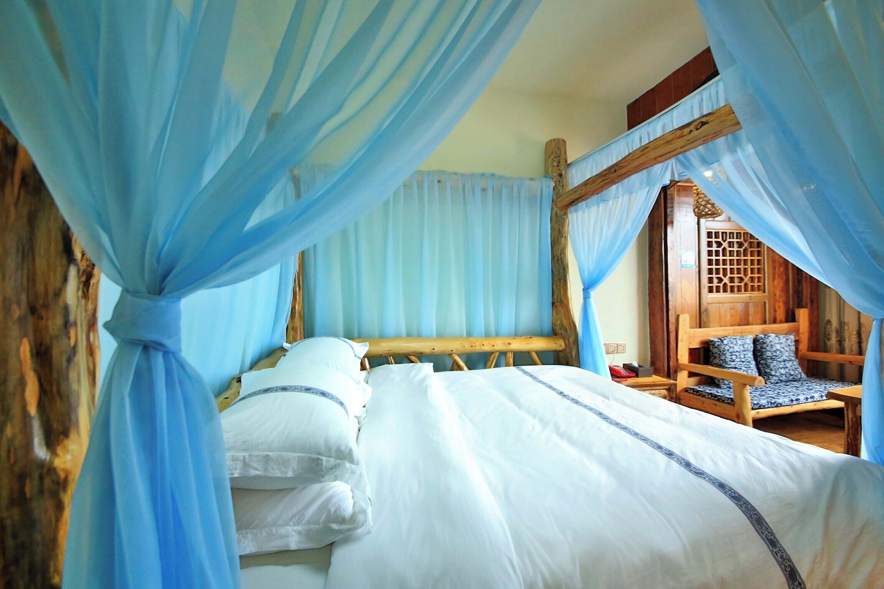 Una cama con ropa de cama blanca y detalles azules, un banco de madera con cojines estampados, y una habitación con cortinas azules y una puerta de madera.