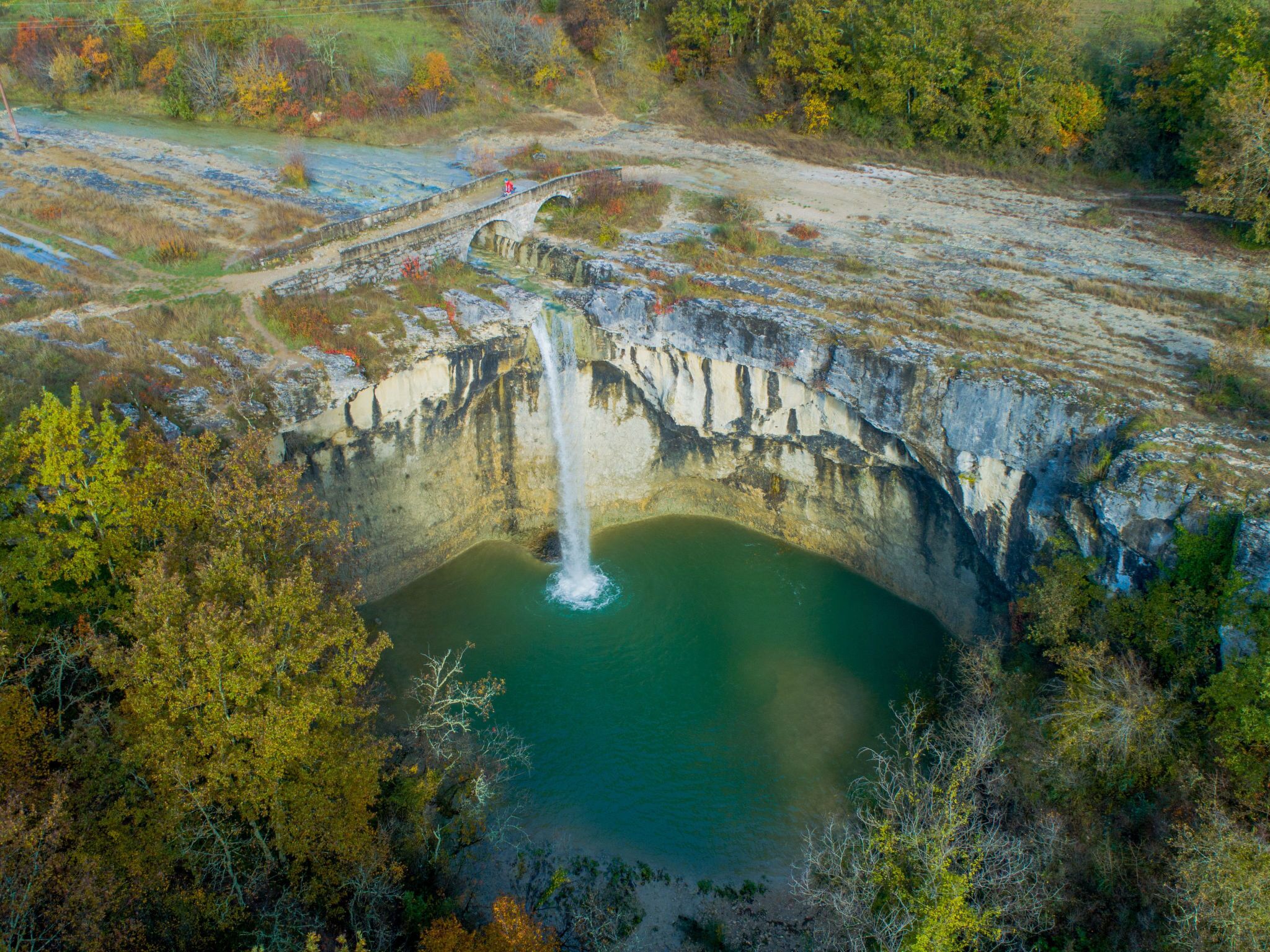 An aerial view of a natural landscape with a waterfall, a bridge, and surrounding vegetation.