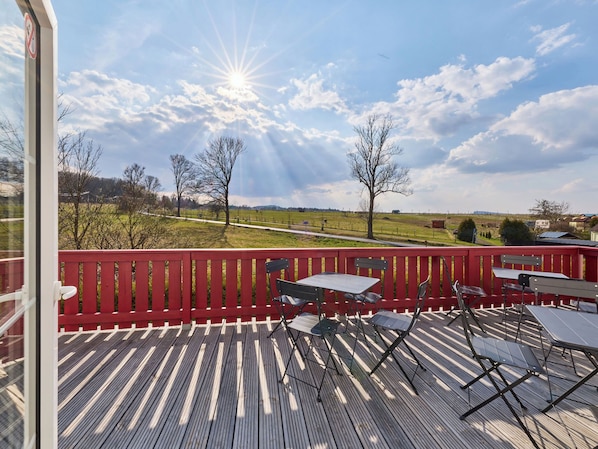 Eine Holzterrasse mit Tischen und Stühlen, ein rotes Geländer und Blick auf eine ländliche Landschaft mit Bäumen und einem Weg.