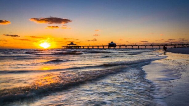 Escena de playa con olas, un muelle y un atardecer.