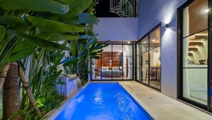 Area piscina esterna moderna con acqua cristallina, circondata da vegetazione lussureggiante e un edificio contemporaneo con ampie vetrate.