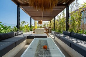 Área externa com mesa de vidro, assentos acolchoados e luminária pendente.