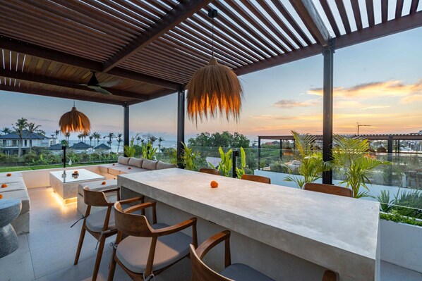 An outdoor dining area with a long table and chairs, surrounded by plants and overlooking a scenic view. - Villa Sevu 2 (Cemagi)