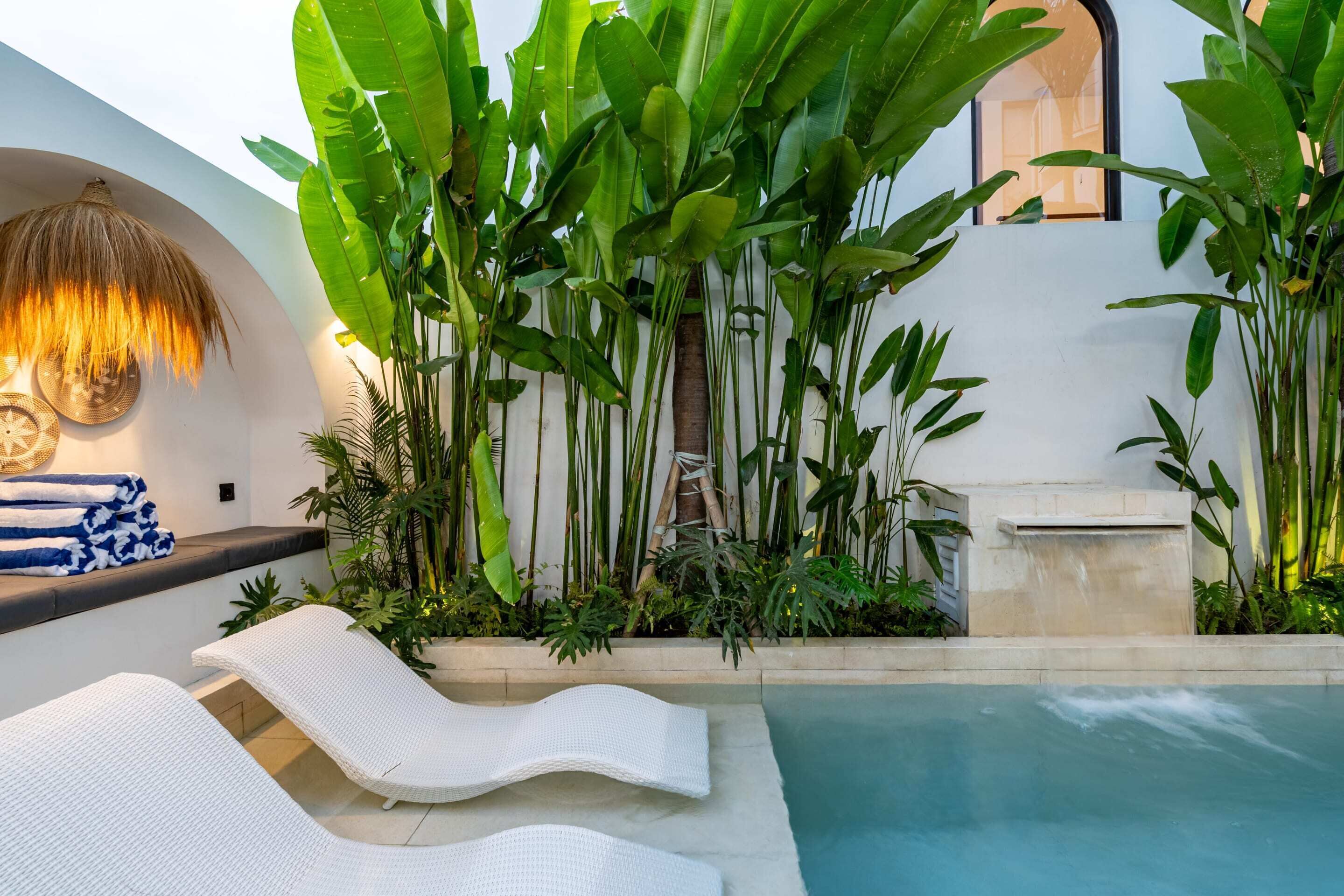 Zona junto a la piscina con sillones de descanso, una lámpara de techo de paja y exuberante vegetación tropical.