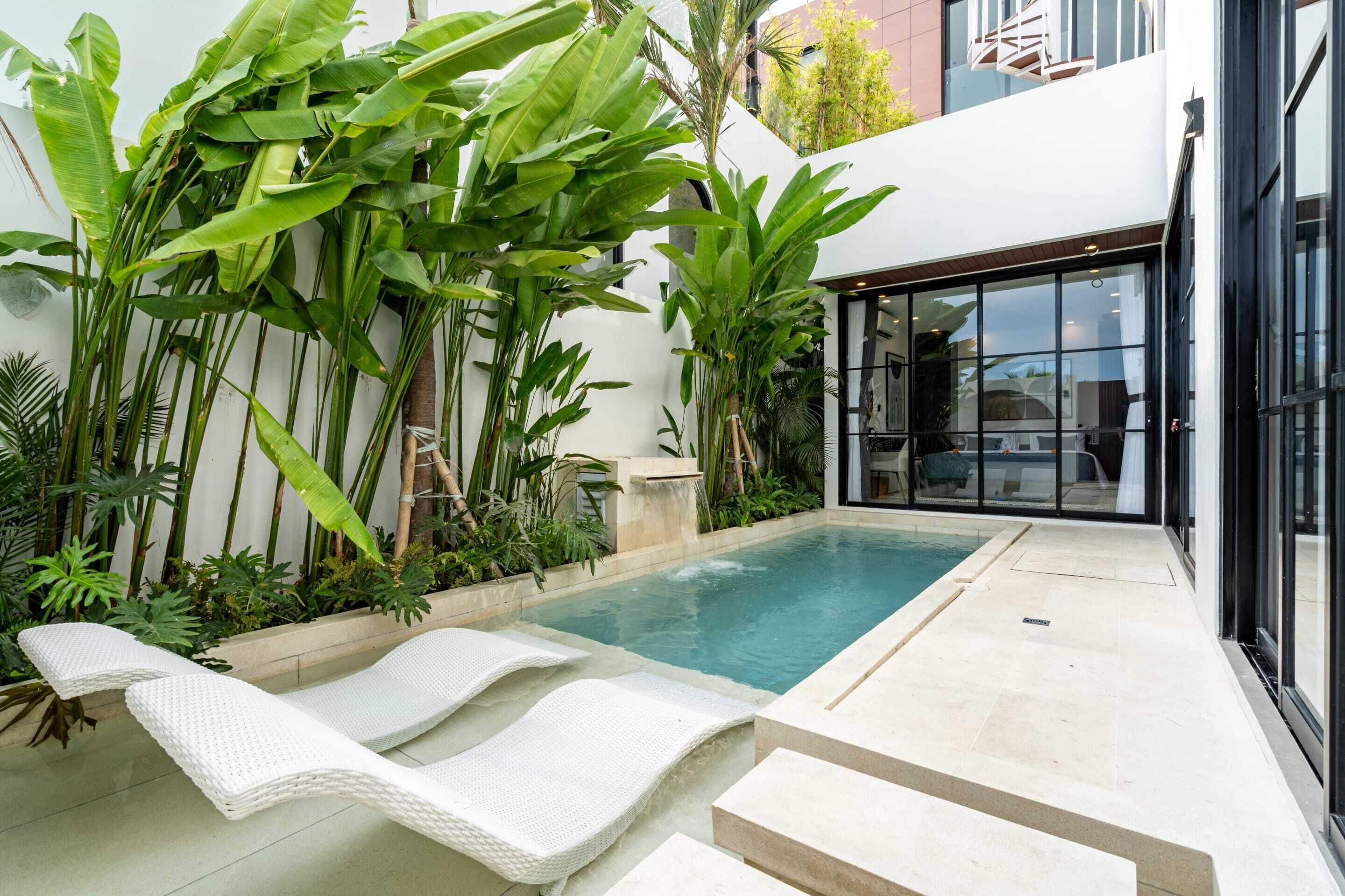 Une piscine entourée de chaises longues blanches, de plantes tropicales luxuriantes et d’un bâtiment moderne doté de grandes fenêtres.