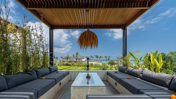 An outdoor seating area with a glass-top table, surrounded by dark cushioned sofas, under a wooden pergola with a hanging light fixture, overlooking a lush garden and a view of a house and ocean. - Villa Sevu 4 (Cemagi)