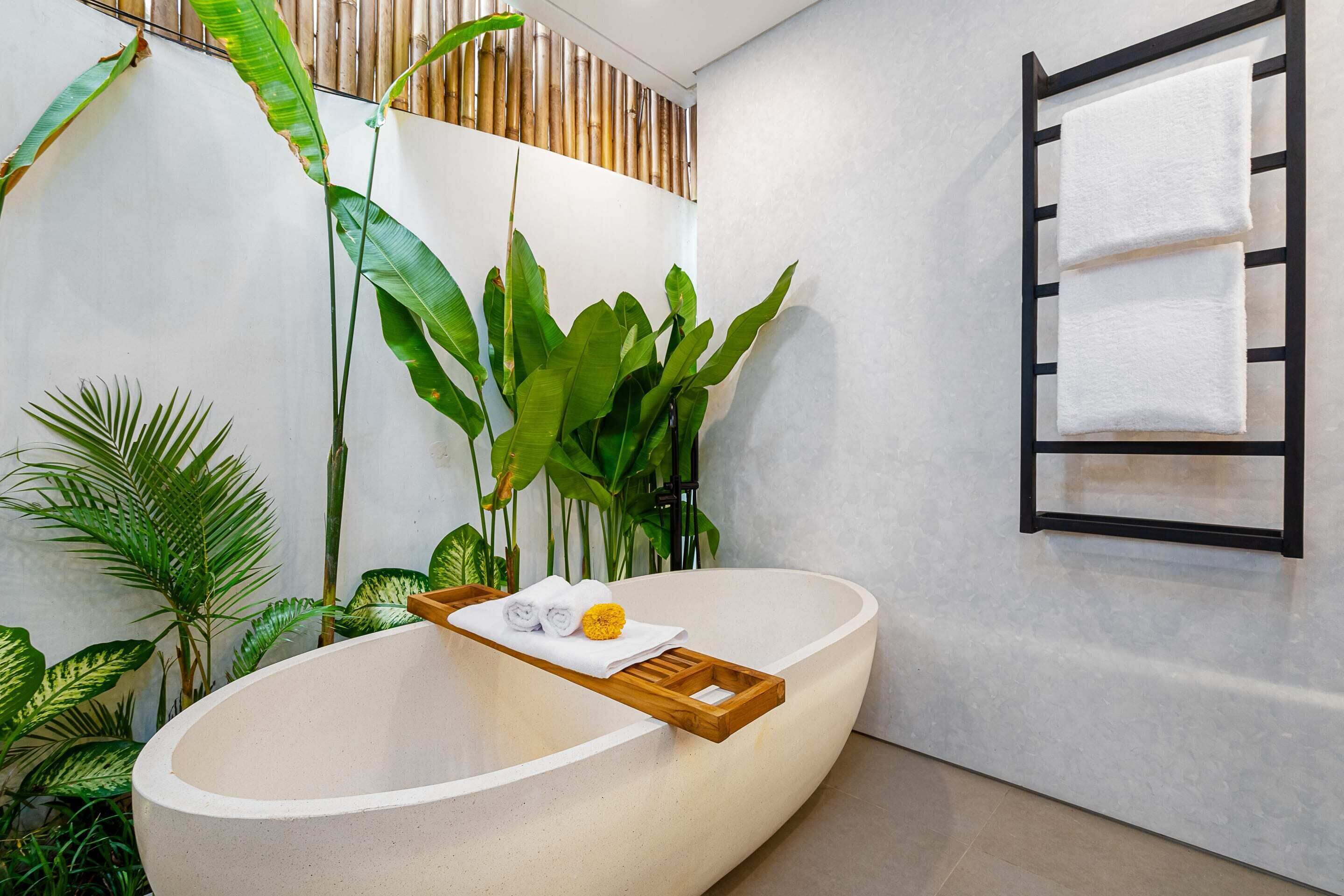 A modern bathroom with a freestanding bathtub, a wooden tray with towels, and lush green plants.
