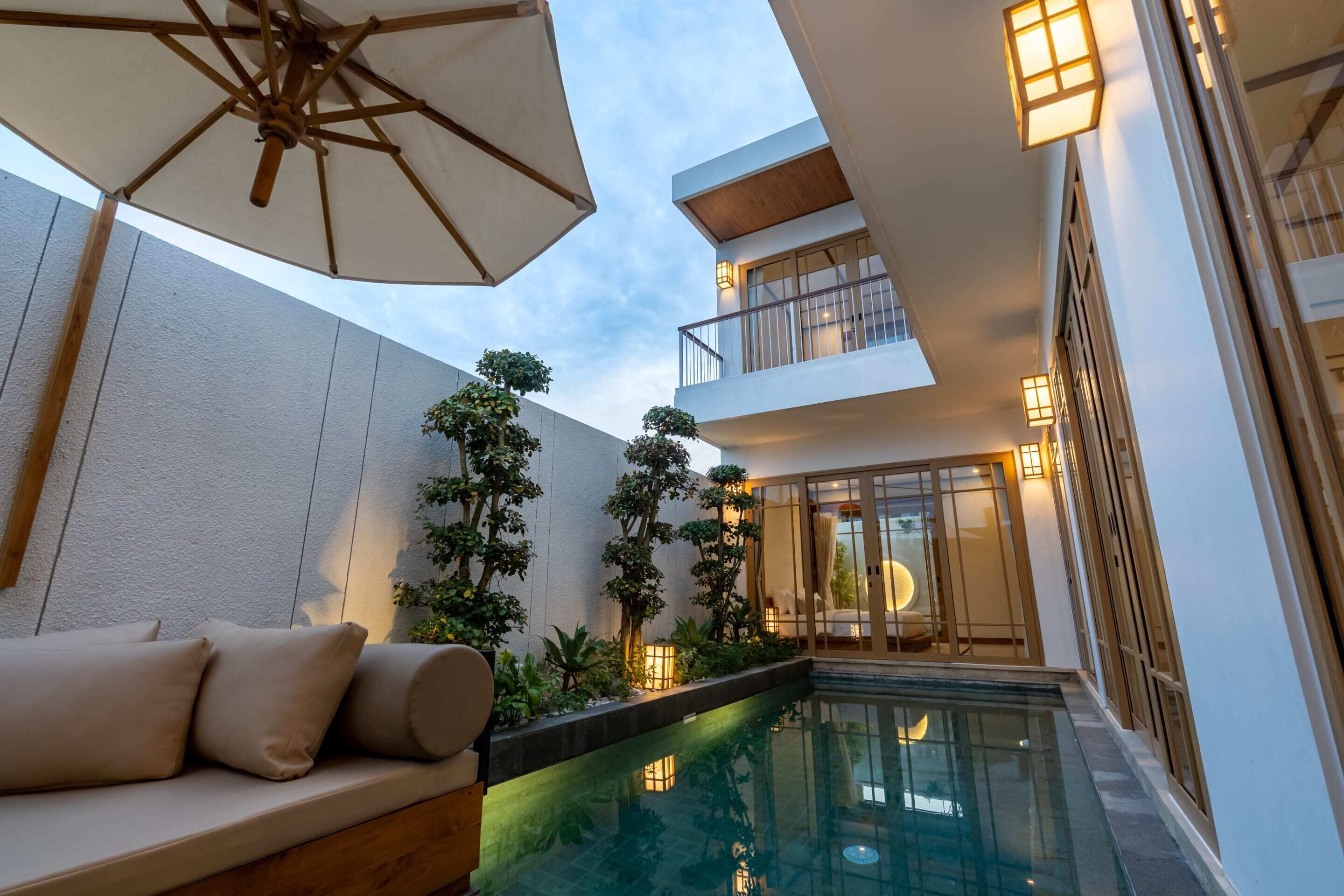 Een modern buitenzwembad met een zithoek, een overdekt terras en een gebouw met balkons en verlichting.