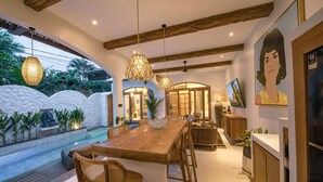 A spacious dining area with a wooden table and chairs, pendant lighting, and a view of a pool and garden. - Villa Jackie O (Seminyak)