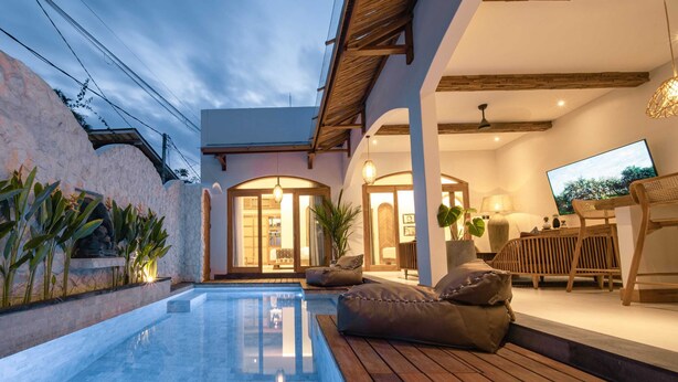 A modern outdoor pool area with a wooden deck, seating, and a view of the interior through a glass door. - Villa Jackie O (Seminyak)