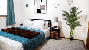 A bedroom with a bed, a nightstand, a lamp, a large potted plant, and abstract wall art.