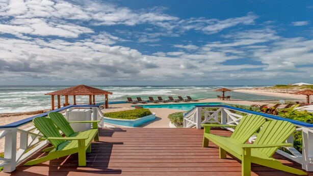 A wooden deck with green Adirondack chairs, a pool, and beach views.