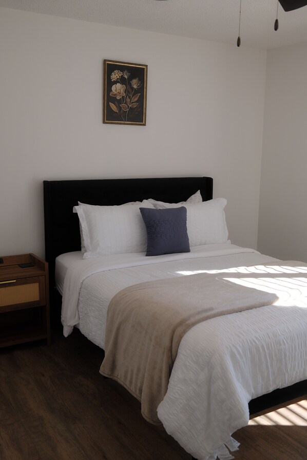 A neatly made bed with white bedding and a dark headboard, a small wooden nightstand beside it, and a framed floral artwork on the wall.