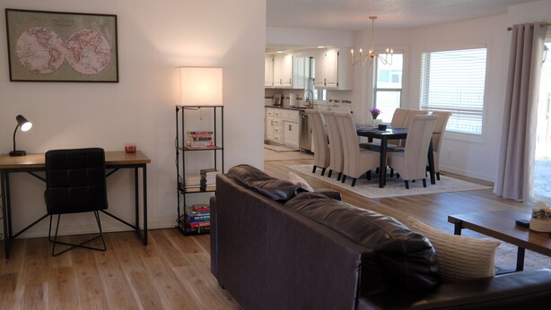 A modern living room with a sofa, a desk with a lamp, and a bookshelf. There is a kitchen in the background with a dining table and chairs. - Comfortable Home Near CoxHealth & MSU - Fenced Yard (Springfield)