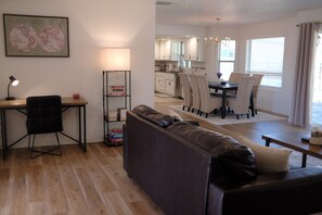 A modern living room with a sofa, a desk with a lamp, and a bookshelf. There is a kitchen in the background with a dining table and chairs.