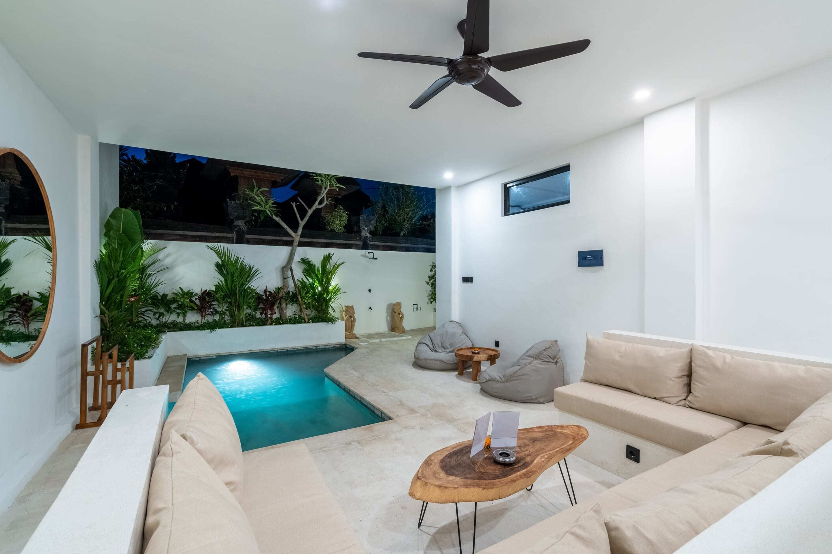 A modern indoor pool area with a ceiling fan, beige sofas, and a small wooden table.
