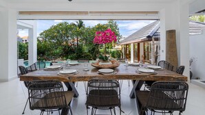 A dining area with a large wooden table set for six, surrounded by wicker chairs, with a view of a pool and greenery outside. - Villa Imbuh 1 (Canggu)