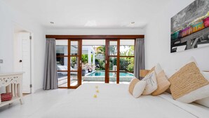 A bedroom with a large bed, a view of a swimming pool, and a balcony with curtains. - Villa Imbuh 1 (Canggu)