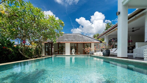 A clear blue pool with a stone statue in the center, surrounded by lush greenery and a white building with a tiled roof. - Villa Imbuh 1 (Canggu)