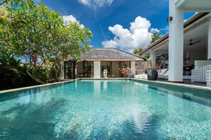 A clear blue pool with a stone statue in the center, surrounded by lush greenery and a white building with a tiled roof. - Villa Imbuh 1 (Canggu)