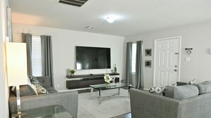 A modern living room with a grey sofa, a glass coffee table, a flat-screen TV, and a white door.
