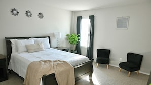 A bedroom with a bed, a nightstand, a lamp, a potted plant, and two chairs.