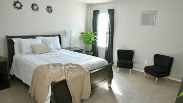 A bedroom with a bed, a nightstand, a lamp, a potted plant, and two chairs.