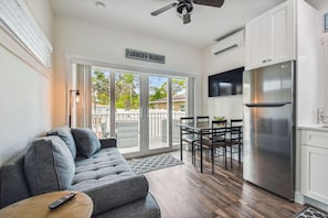 A modern kitchen with a stainless steel refrigerator, a dining area with a table and chairs, and a grey sofa with a remote control on a side table. - Dunedin Tiny House Combo/Sleeps 16! Walk or Bike to Downtown Dunedin! (Dunedin)