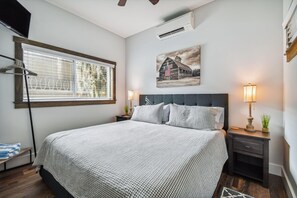 Et pænt opredt soveværelse med en stor seng, et sengebord med lampe, en lille plante og et vindue med persienner.