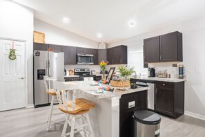 A modern kitchen with dark cabinets, a stainless steel refrigerator, a microwave, and a dishwasher. There are two white bar stools and a small island with a sink.