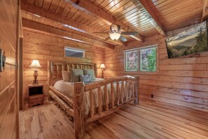 A wooden cabin bedroom with a bed, two bedside lamps, a ceiling fan, and a window with a view of trees. - Charming Gatlinburg cabin for two with stunning mountain views. (Gatlinburg)