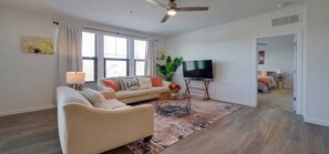 A modern living room with a sofa, a coffee table, a flat-screen TV, and a large window with blinds. - Inviting 2 BR • Near IMG Academy • Pool  (Bradenton)