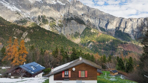 Un chalet doté d’un toit en pente, d’une façade en bois et d’un petit balcon. Il est entouré d’un paysage enneigé, avec des montagnes en arrière-plan.