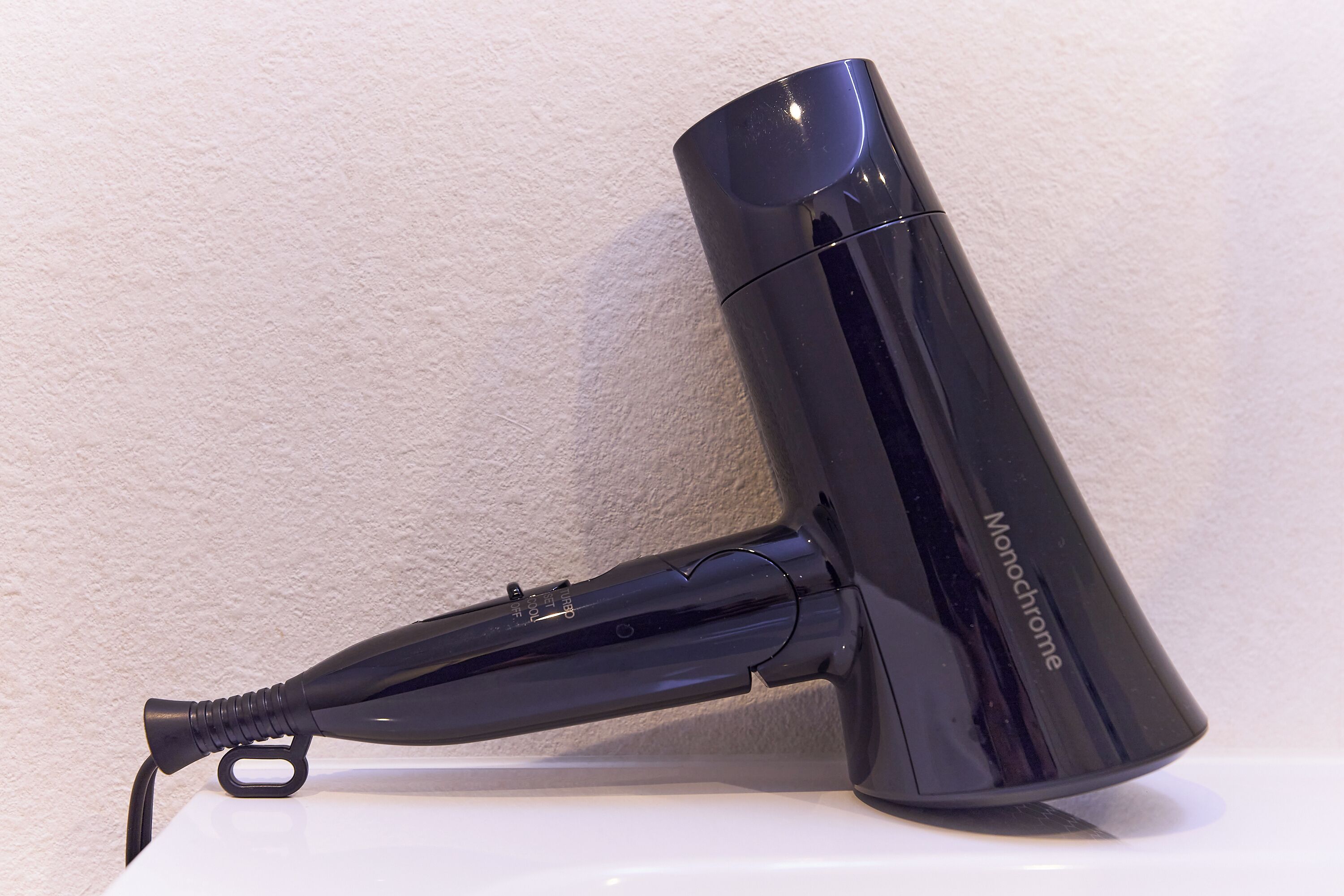 A black Monochrome hair dryer with a cord and a power adapter on a white surface against a textured wall.