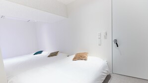 A single bed with white bedding and two pillows, a white door with a handle, and a light switch on the wall. - VORT Yamabukicho Residence (Tokyo)