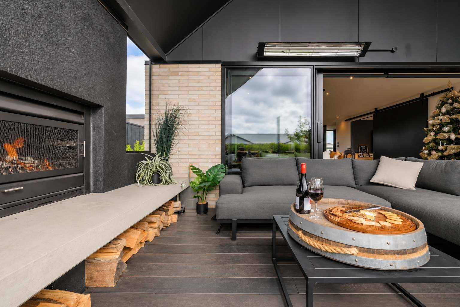 A modern outdoor seating area with a built-in fireplace, a round wooden table with food and drink, and a grey sofa.