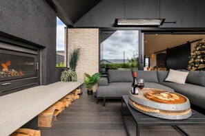 A modern outdoor seating area with a built-in fireplace, a round wooden table with food and drink, and a grey sofa.