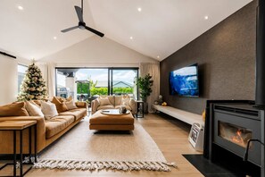 A modern living room with a sofa set, a coffee table, a fireplace, and a large TV on the wall. - Stylish Home | Private Spa + Indoor & Outdoor Fire (Prebbleton)