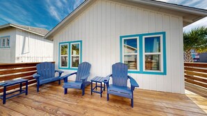 Una terraza de madera con sillas Adirondack y una pequeña mesa, un edificio blanco con detalles azules y ventanas, y un cielo azul despejado.