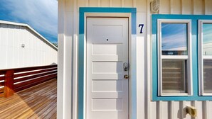 Una puerta blanca con marco azul y el número '7' encima, una terraza de madera y una ventana con marco azul.