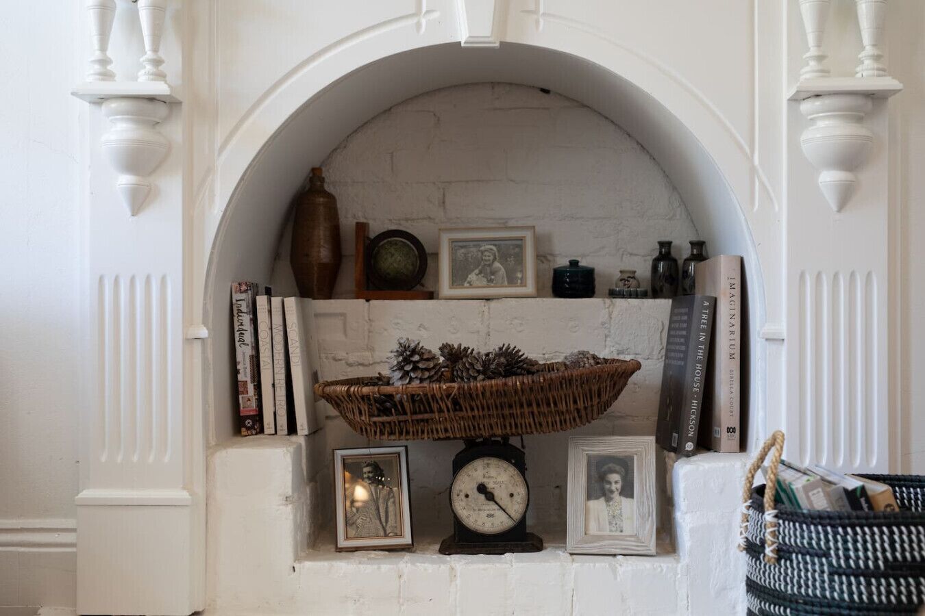Lareira com manto branco, rodeada por livros, fotografias emolduradas e um cesto decorativo.