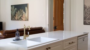 A kitchen with a white countertop, a gold faucet, and a dishwasher. There is a brown leather couch in the background and a door leading to another room. - The Countess Central Heritage in Launceston (Launceston)