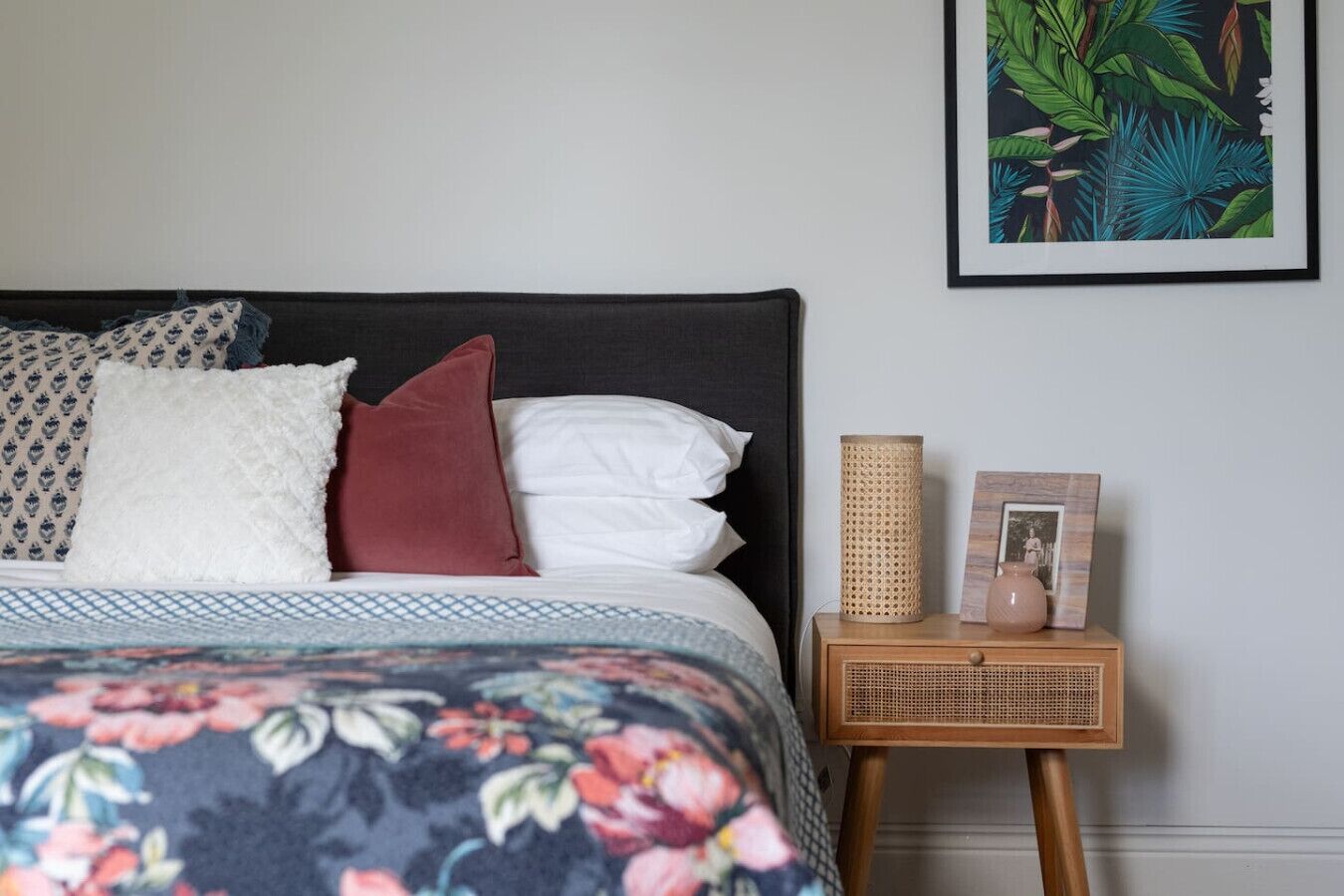 A neatly made bed with a floral duvet, a dark headboard, and a bedside table with a lamp and picture frame.