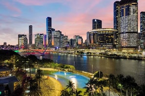 A city skyline with modern high-rise buildings, a river, and a well-lit swimming pool area with palm trees. - City Beach At your door! 3BR retreat at South Bank (South Brisbane)