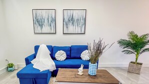 Ein Wohnzimmer mit einem blauen Sofa, einem weißen Pouf, einem Holztisch und einem weißen Teppich. An den Wänden befinden sich zwei abstrakte Gemälde und ein Blumentopf.