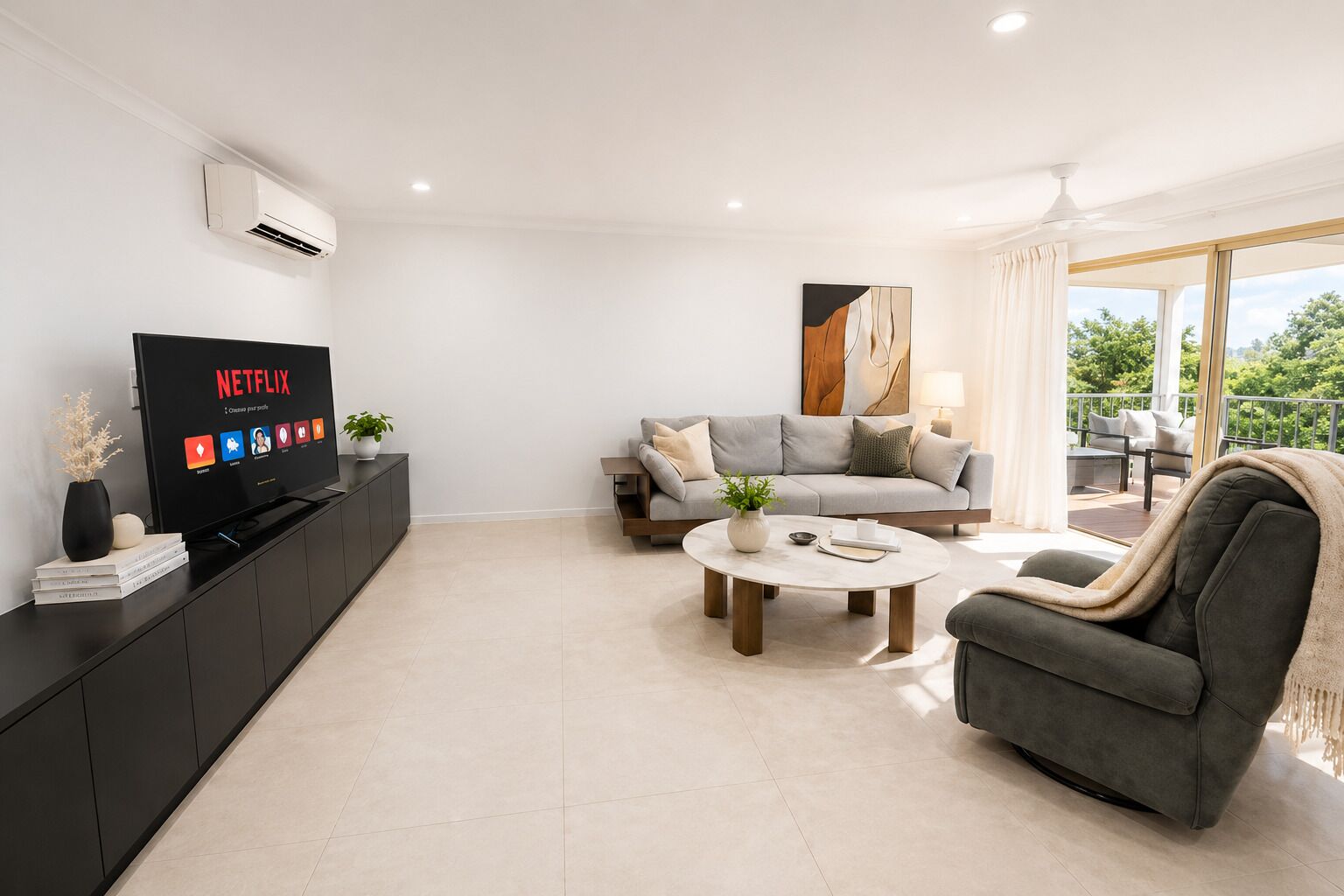A modern living room with a flat-screen TV, a grey sofa, a coffee table, and a balcony with a view.