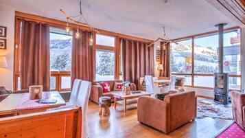 Ein gemütliches Wohnzimmer mit Kamin, Holzmöbeln und großen Fenstern mit Blick auf verschneite Berge.